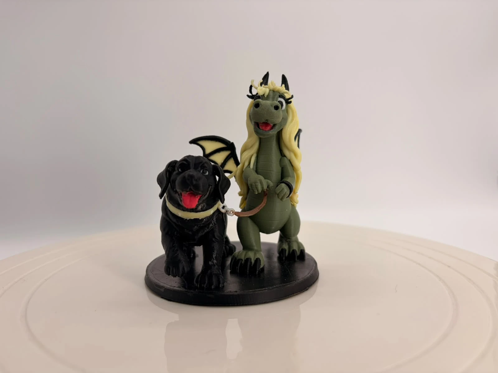 3D-gedruckter grüner Drache mit blonden Haaren und Flügeln, der einen schwarzen Hund an der Leine führt, auf rundem Sockel